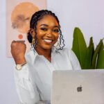 Smiling businesswoman works confidently on a laptop in a creative office setting, embodying modern entrepreneurship.